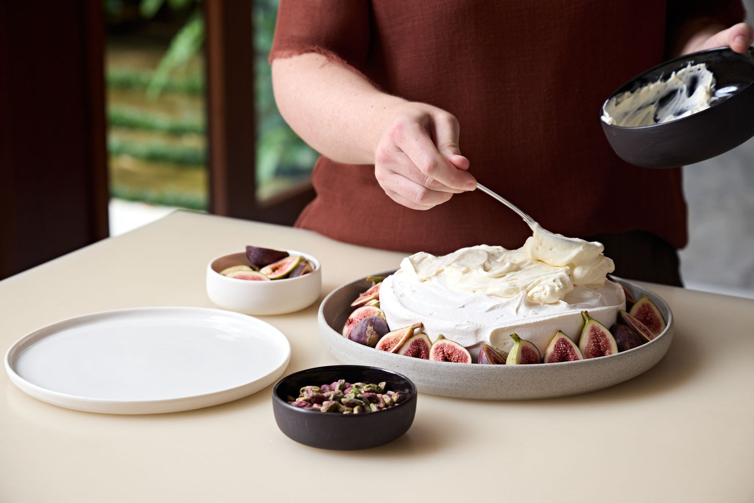 The image shows Sophie covering a pavlova in cream, figs and pistachios. The pavlova sits in a grey platter. A dip bowl of figs and another of pistachios sits on the bench.